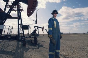A man in overalls and hard hat at a pump jack in open ground at an oil extraction site.